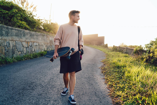 Full length of happy young male skater in casual outfit in back lit carrying skateboard while walking along asphalt road in countryside in sunset lights - Foto, afbeelding