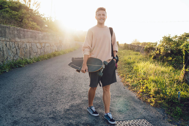Half length portrait of cheerful caucasian male skater walking on rural asphalt road enjoying summer weekends, handsome hipster guy 20s with board spending free time on active sport hobby outdoors - 写真・画像