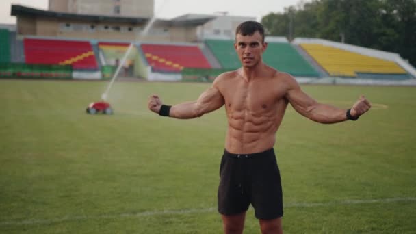Een bodybuilder staat onder druppels water van een sprinkler op een voetbalveld en toont spieren. Vrijheidsconcept. Langzame beweging - Video