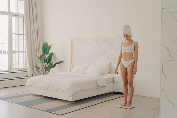 Снимок здоровой молодой худой женщины в нижнем белье, стоящей на весах в спальне - Фото, изображение