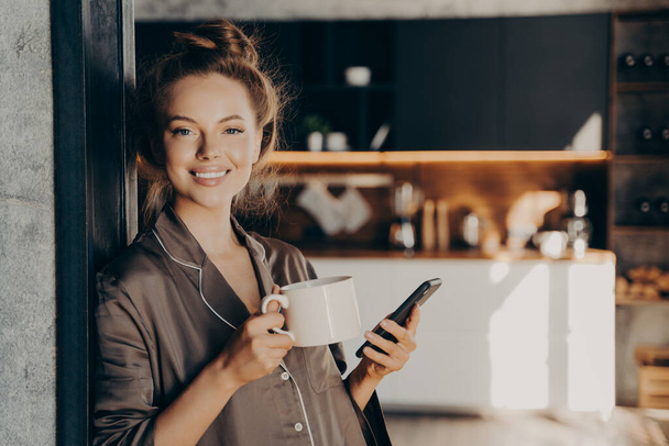 Piękny szczęśliwy młoda kobieta o jej poranną kawę podczas sprawdzania nowych e-maili i powiadomień na smartfonie - Zdjęcie, obraz
