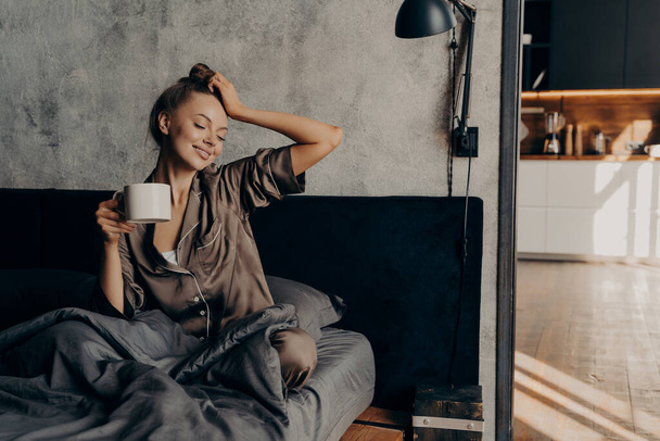 Muotokuva nuori rento nainen tyylikäs kotipuku herääminen kuppi kahvia sängyssä - Valokuva, kuva
