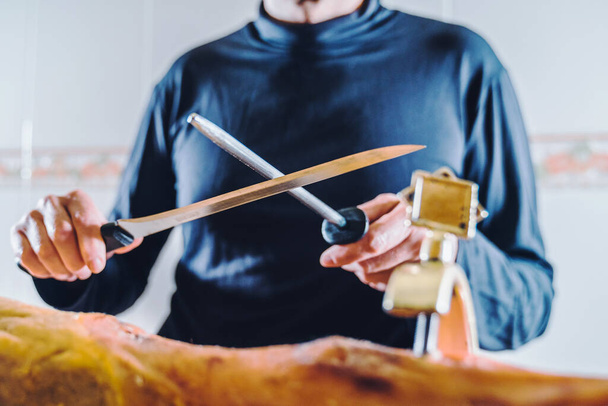 Anonymní člověk brousí šunkový nůž před krájením šunky. Velmi dobře nabroušený nůž. Střední část ostrého nože šéfkuchaře v kuchyni. - Fotografie, Obrázek