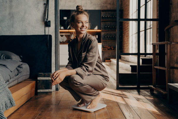 Image d'un joyeux jeune mannequin en pyjama satiné accroupi sur des échelles avec un large sourire dans la chambre moderne - Photo, image