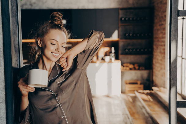 Joyeux belle brune femelle stratching avec une tasse de café dans sa main tout en se tenant dans la cuisine - Photo, image