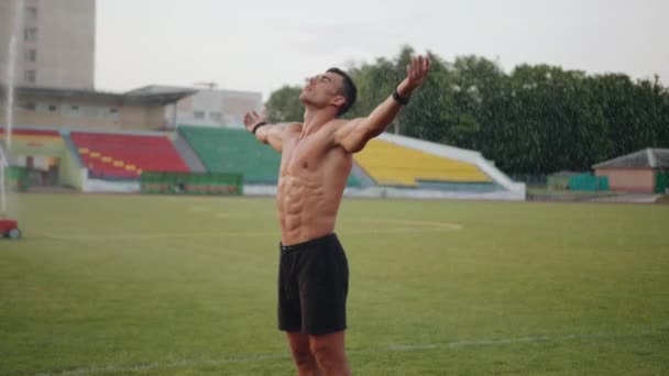 Un hombre musculoso está bajo gotas de agua de un aspersor en un campo de fútbol con los brazos extendidos. En cámara lenta. Concepto de libertad y éxito - Imágenes, Vídeo