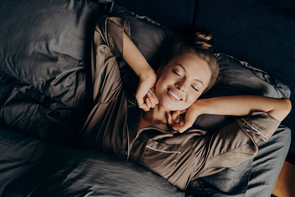 Νεαρή όμορφη χαλαρή μελαχρινή γυναίκα σε σατέν πυτζάμα τέντωμα στο κρεβάτι - Φωτογραφία, εικόνα