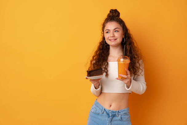 Young fit curly woman holding a piece of cake against yellow background - Zdjęcie, obraz