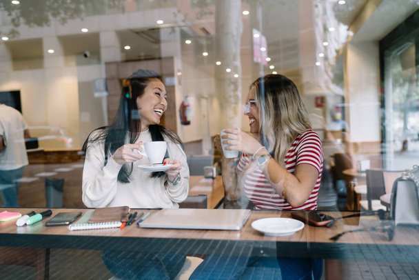 Cheerful multiracial female best friends laughing telling funny stories during conversation in cafe interior, smiling young women enjoying meeting on lunch break drinking coffee and communicating - Φωτογραφία, εικόνα