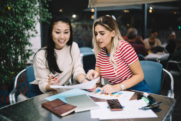 Successful hipster girls talking during cooperation meeting for checking informative documents in street cafe, diverse female planning organisation while doing university homework and course work - 写真・画像