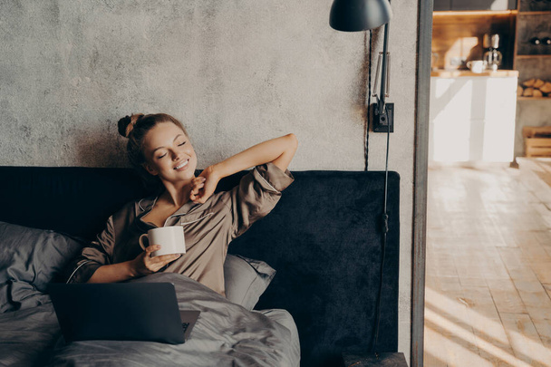 Happy beautiful female with closed eyes drinking coffee in bed before remote work at home - Foto, Imagem