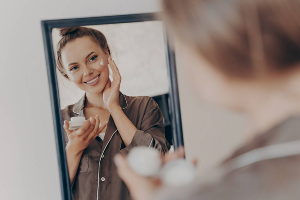 Reflectie in spiegel van mooie vrouw in zijde pyjama glimlachen tijdens het aanbrengen van gezichtscrème voor diepere reparatie - Foto, afbeelding