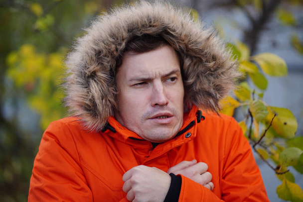 Portrait of adult man in hood and orange jacket cringing from cold in autumn season. Funny male froze while standing on street in cold weather - Фото, зображення