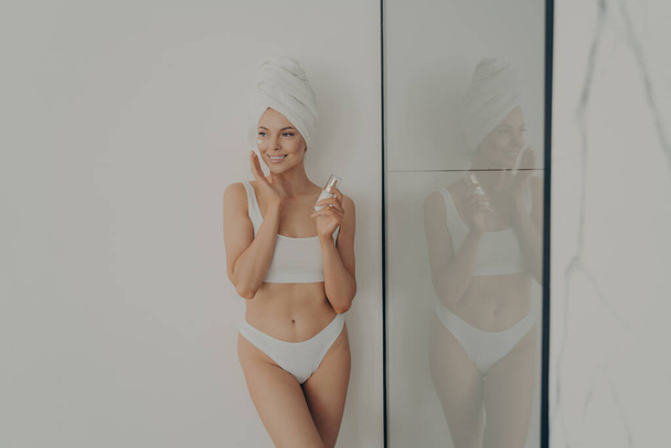Bella donna in piedi in bagno dopo la doccia del mattino e l'applicazione di crema idratante sul viso - Foto, immagini