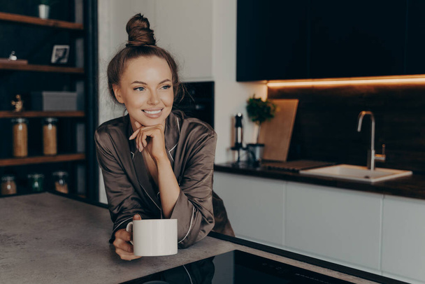 Entspannte positive junge Frau im seidenen Pyjama bei einer Tasse Kaffee im stilvollen Kücheninterieur - Foto, Bild