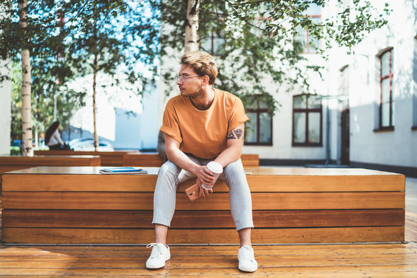 Serious man in glasses and stylish clothes holding disposable cup of fresh coffee and looking away while chilling on wooden bench - Foto, immagini