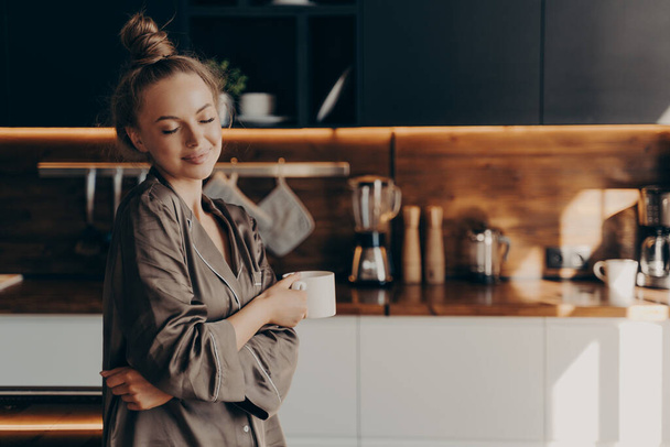 Όμορφη νεαρή ευτυχισμένη γυναίκα στέκεται σε ζεστό μεταξωτές πιτζάμες στην κουζίνα με φλιτζάνι καφέ - Φωτογραφία, εικόνα