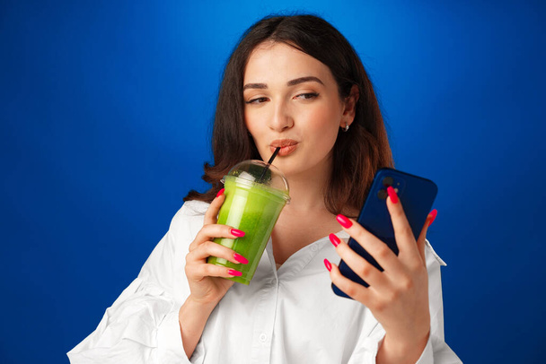 Young attractive happy woman holding a cup of green smoothie and using her smartphone against blue background - Foto, Imagen