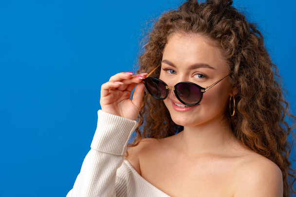 Retrato de una joven mujer de pelo rizado con gafas de sol sobre fondo azul - Foto, Imagen