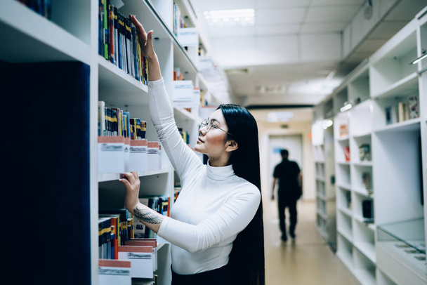 Side view of Asian female reader with tattoo on hand standing in front of bookshelf and searching for book on top shelf - Фото, изображение