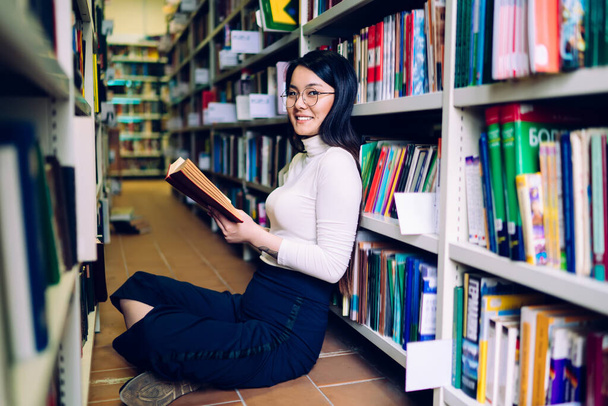 Dunkelhaarige asiatische fröhliche Frau mit Brille sitzt im Schneidersitz neben Bücherregal mit aufgeschlagenem Buch in der Hand und blickt in die Kamera - Foto, Bild