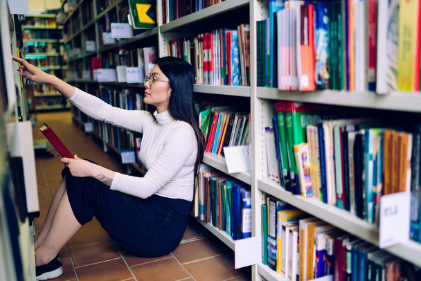 Side view of Asian female student sitting on floor with book in hand leaning on bookshelf and searching for literature in university library - Foto, Imagem
