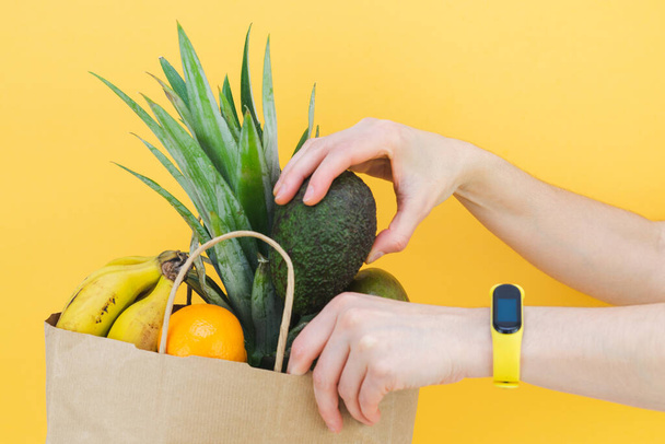 Woman hands filling paper bag with assorted fruit on yellow background. Copy space. - Photo, Image