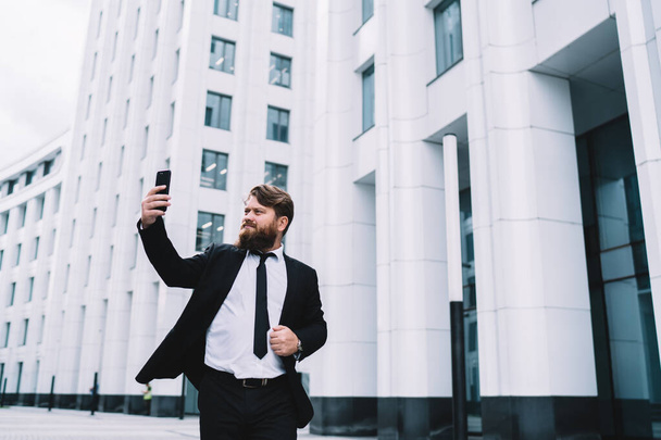 Serious man in formal shirt and jacket with beard using smartphone for taking selfie while standing in front of big office building - Photo, Image
