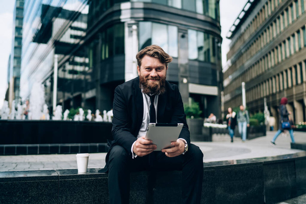 Positive bearded businessman in formal clothing and tie sitting on stone border and looking at camera while surfing tablet outside - Φωτογραφία, εικόνα
