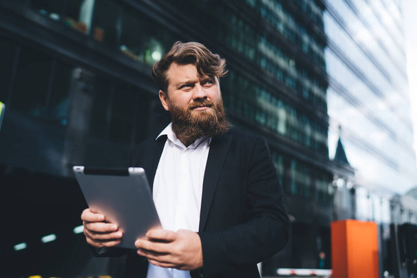 Concentrated and confident businessman with tablet in white shirt and black jacket looking away while standing against tower with glass walls - Valokuva, kuva