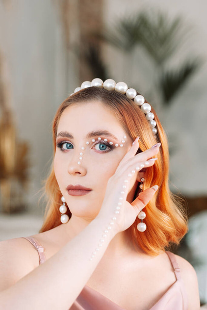 fashion portrait of a woman with an orange curly square and with makeup made of mother-of-pearl beads glued to her face and hand, with long pearl earrings and a headband on her head - Foto, Bild