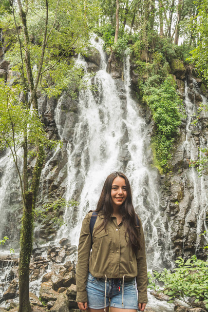 Chica aventurera explorando cascadas en medio de la selva, con ropa ligera encuentra paisajes increíbles. Lleva un cortavientos verde y pantalones cortos de mezclilla azul.. - Foto, Imagen
