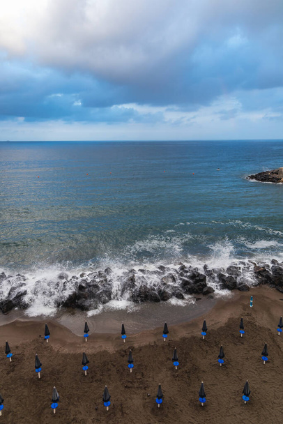 Velké mořské vlny narážejí na skály vedle pláže plné deštníků - Fotografie, Obrázek