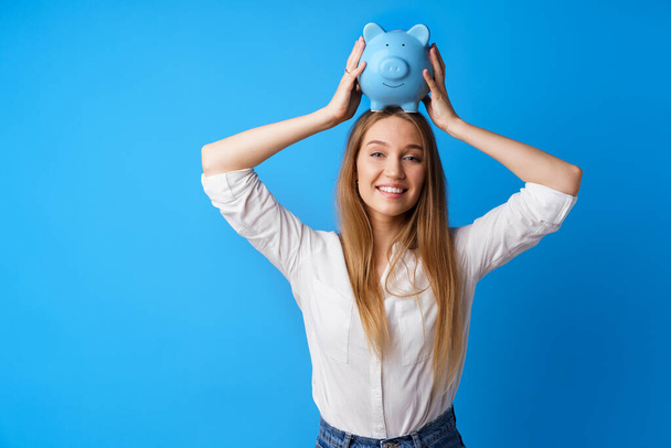 Beautiful smiling young woman with piggy bank on blue background - Valokuva, kuva