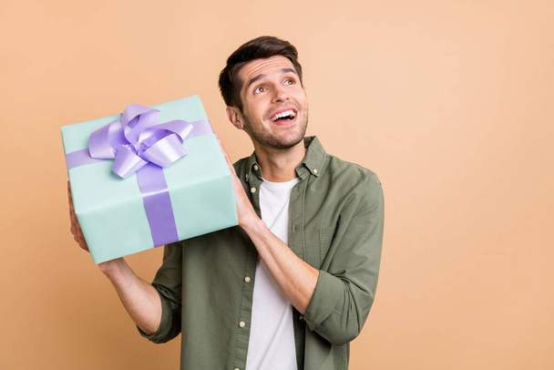 Photo of amazed dreamy young happy man look empty space hold gift box smile isolated on beige color background - Photo, image
