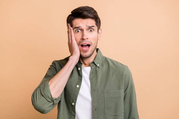 Photo of tired shocked young guy dressed green shirt arm cheek open mouth isolated beige color background - Photo, Image