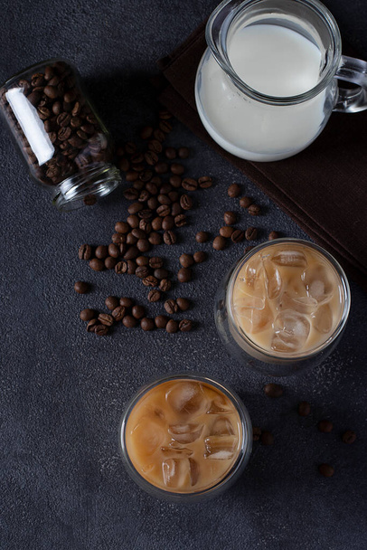 Two glasses of cold coffee with ice cubes, coffee beans and jug of milk on a dark background. Top view - Photo, Image