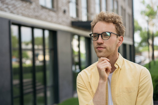 Retrato de un hombre guapo pensativo con camisa amarilla y gafas con estilo que planea comenzar mirando hacia otro lado en el espacio de copia, de pie en la calle - Foto, Imagen