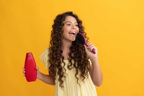 счастливая девушка-подросток с длинными вьющимися волосами держать бутылку шампуня поет в расческе, весело - Фото, изображение