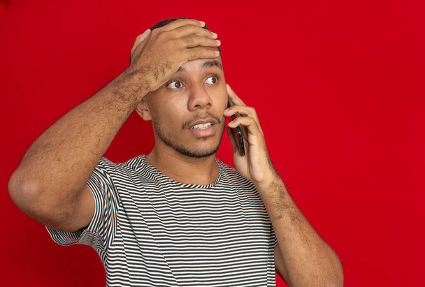 Ένας άντρας που χρησιμοποιεί κινητό κάνει μια ανήσυχη χειρονομία με κόκκινο φόντο. - Φωτογραφία, εικόνα