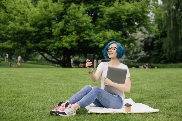 Mavi saçlı genç kadınlar ellerinde akıllı telefonlar ve dizüstü bilgisayarlarıyla yeşil çimlerde oturuyorlar. Yaz zamanı. - Fotoğraf, Görsel