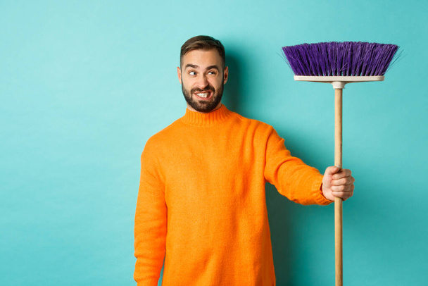 Φίλος ψάχνει σύγχυση σε σκούπα, αναγκαστική καθαρό σπίτι, κάνει δουλειές, στέκεται δυσαρεστημένος με ανοιχτό μπλε φόντο - Φωτογραφία, εικόνα