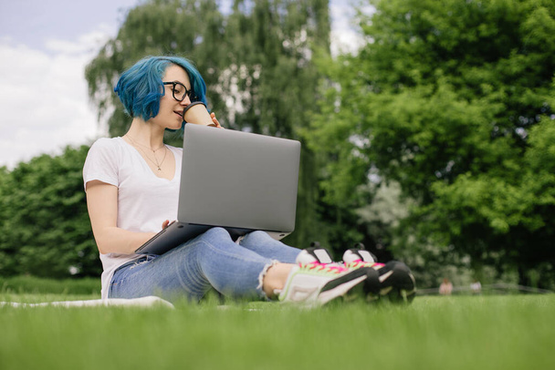 Mujeres jóvenes con el pelo azul sentado en la hierba verde con el ordenador portátil y el café en las manos. Hora de verano - Foto, imagen