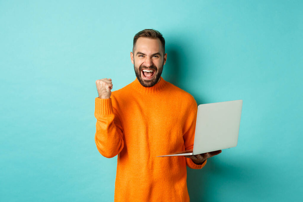 Freelance and technology concept. Lucky man winner celebrating, winning online, showing fist pump and holding laptop, standing over light blue background - Valokuva, kuva