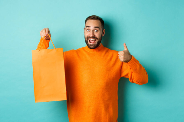 Uomo felice che tiene la shopping bag arancione e gioisce dello sconto e celebra, mostrando i pollici in alto e raccomandando, in piedi sopra lo sfondo blu - Foto, immagini