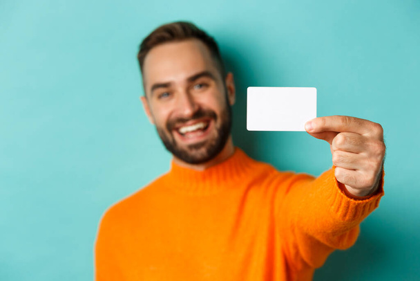 Primer plano del hombre caucásico guapo que va de compras, mostrando la tarjeta de crédito y sonriendo, de pie sobre fondo turquesa - Foto, imagen