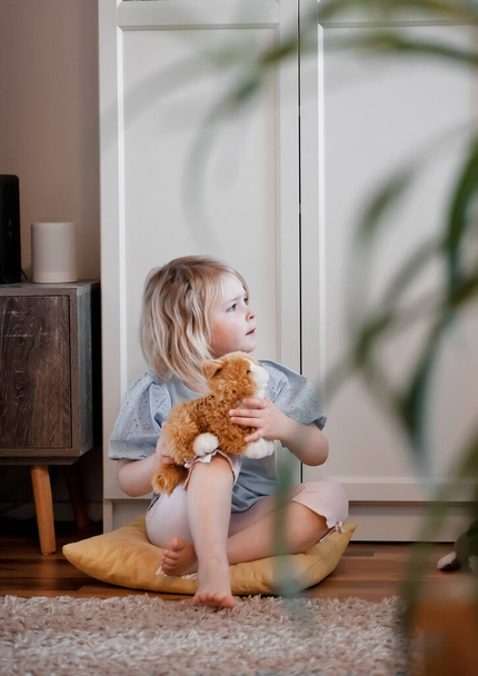 Mała dziewczynka siedząca na podłodze w salonie i bawiąca się kotem zabawki, strzał wewnątrz, naturalne światło - Zdjęcie, obraz