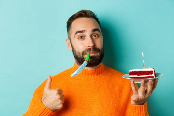 Κοντινό πλάνο του αστείο ενήλικας γιορτάζει τα γενέθλιά του, κρατώντας τούρτα bday με κερί, blowing κόμμα wistle και δείχνει τον αντίχειρα-up, στέκεται πάνω από το γαλάζιο φόντο - Φωτογραφία, εικόνα