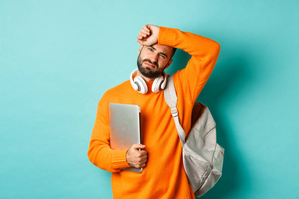 Cansado estudiante masculino limpiando el sudor de la frente, sosteniendo el ordenador portátil y la mochila, de pie en suéter naranja por fondo turquesa - Foto, imagen