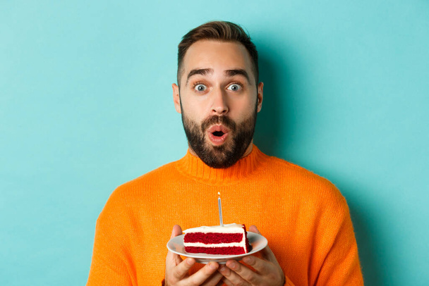 Nahaufnahme eines glücklichen erwachsenen Mannes, der Geburtstag feiert, Kuchen mit Kerze hält und Wünsche äußert, vor türkisfarbenem Hintergrund steht - Foto, Bild